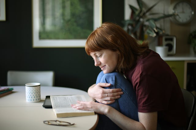 A person with red hair sits at a round table, cradling a book and smiling softly. A mug and plant ar...